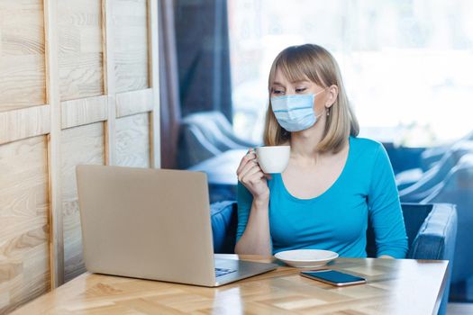 Portrait of young woman with surgical medical mask in blue t-shirt is sitting and working and looking at laptop screen and drinking coffee. Indoor distance working, medicine and health care concept.