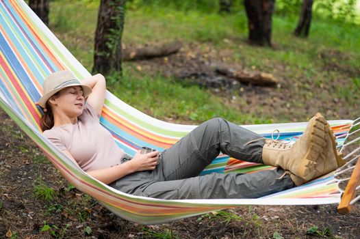 Caucasian woman drinks hot tea from a thermo mug while lying in a hammock in nature