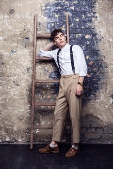 full length fashionable man in white shirt and beige pants with suspenders posing near old wooden ladder on brick wall background, standing and looking away. indoor, studio shot.