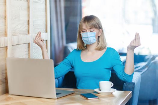 Portrait of calm young blonde woman with surgical medical mask in blue t-shirt is sitting and working on laptop and try to relax in yoga pose. Indoor working, medicine and health care concept.