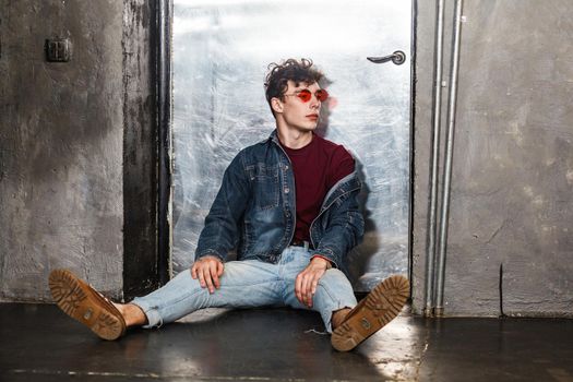 Stylish young fashion model man in bright red sunglasses and denim casual style sitting on floor and posing near metallic door and looking away. indoor, studio shot.