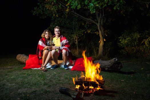 Young happy loving couple sitting on log near the fire covered with plaid holding wine, drinking and enjoy their romantic night. looking at camera with smile.
