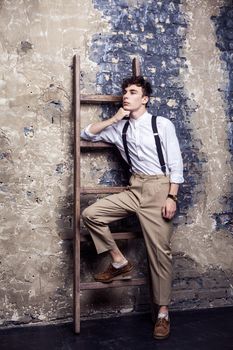 full length fashionable man in white shirt and beige pants with suspenders posing and lean on old wooden ladder on brick wall background. indoor, studio shot.