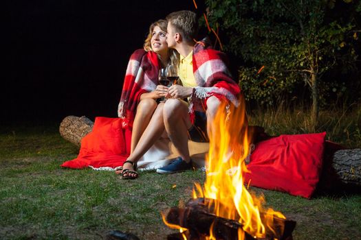 Young happy loving couple sitting on log near the fire covered with plaid holding wine, drinking and enjoy their romantic night. he is kissing her and smiling.