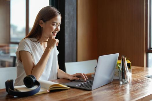 Pensive young asian woman in headphones work on laptop talk speak on video webcam call online. Smart ethnic female in earphones study distant on computer at home. Education concept