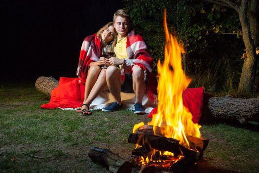Young happy loving couple sitting on log near the fire covered with plaid holding wine, drinking and enjoy their romantic night. hugging and relaxing together.