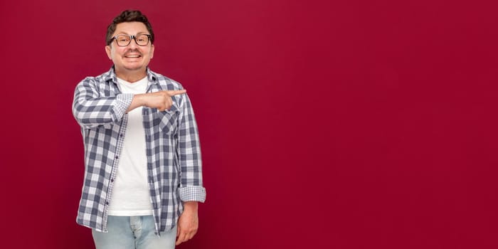 Portrait of happy handsome middle aged business man in casual checkered shirt and eyeglasses standing and pointing at copyspace, looking and toothy smile. studio shot, isolated on dark red background.