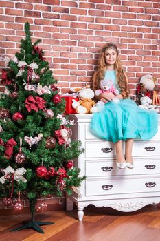 Beautiful teenage blonde sit on a white nightstand near christmas tree, with many toys, toothy smile and looking at camera. Studio shot