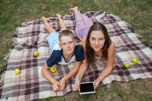 Young sister and brother with freckles on their faces lying down on plaid and using tablet in park. top view, looking at camera with smile.