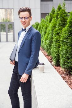 Attractive young businessman with laptop and coffee in hands on office building background. holding laptop and coffee and looking at camera with toothy smile.