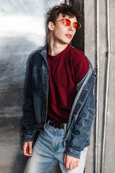 Stylish young fashion model man in bright red sunglasses and denim casual style posing near metallic door, lean and looking at camera with serious face. indoor, studio shot.