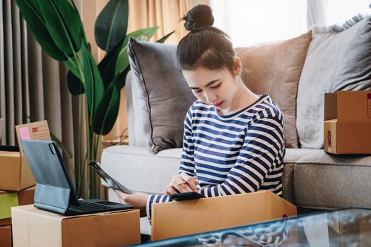 Online selling business idea, beautiful girl checking the products that customers ordered on their mobile phones and tablet in order to pack the products into parcel boxes to deliver to customers