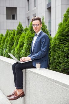 Attractive young businessman with laptop and coffee in hands on office building background. holding laptop and looking at camera with toothy smile.