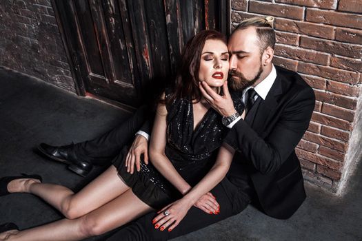 Handsome bearded man sitting on the floor, touching chin woman, while his ginger girlfriend is leaning on him. Studio shot, vintage inerior