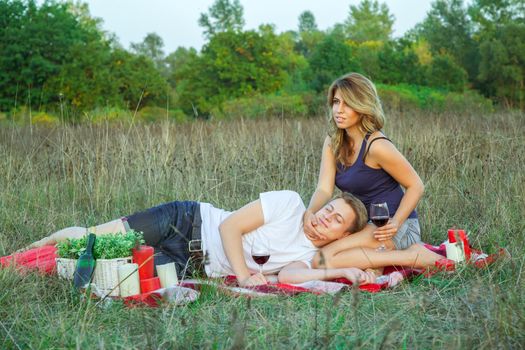 Beautiful young happy loving couple on picnic lying down on plaid in field on sunny summer day enjoying, holding and drinking wine and resting.