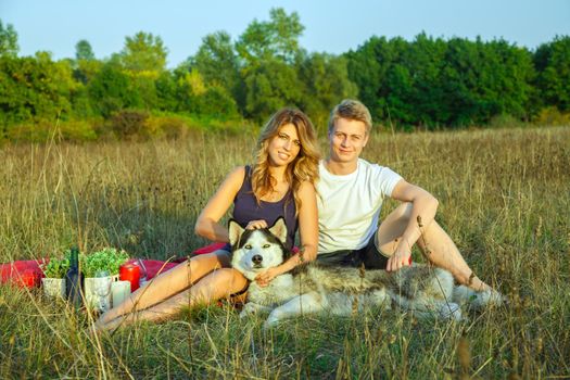 Beautiful young happy loving couple on picnic lying down on plaid with their dog in field on sunny summer day enjoying and resting. looking at camera and smiling.