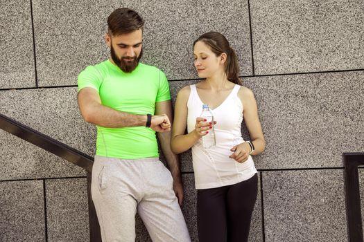 Young sporty couple are resting and check their fitness watch together, drinking water. sport and technology concepts.