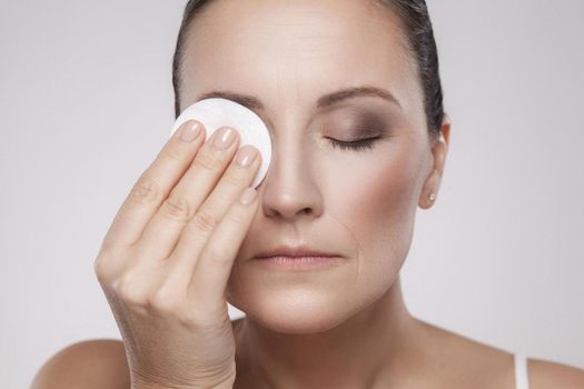 Excited cheerful confident lovely beautiful mature woman is using cotton pad with micellar water for removing make up from eye, isolated on grey background, copy space, studio shot, indoor, close up