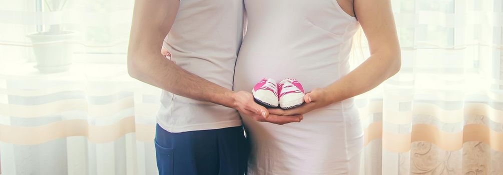 Pregnant woman and man hug belly and hold baby shoes. Selective focus. People.