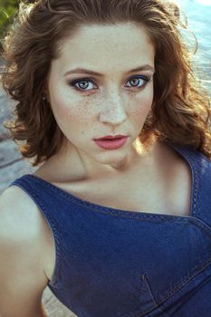 Beautiful young fashion model with freckles on her face and denim blue dress and fashion makeup and hairstyle is lying down on pier, posing and looking at camera.