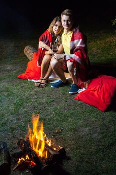 Young happy loving couple sitting on log near the fire covered with plaid holding wine, drinking and enjoy their romantic night. looking at camera and smiling.