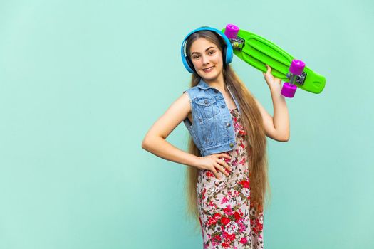 The happy beautiful long haired blonde girl in dress and blue headphones, having fun with green plastic penny skateboard in crossed hands onder head, front of the light blue wall. Isolated studio shot.