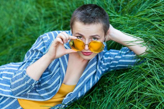 Portrait of serious cute young woman with short hair in casual blue striped suit, yellow shirt and eyeglasses lying down on green grass and looking at camera through glasses. outdoor summertime shot.