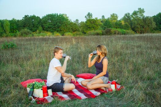 Beautiful young happy loving couple on picnic lying down on plaid in field on sunny summer day enjoying, holding and drinking wine and resting. looking at camera and smiling.