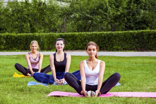 Groupe of beautiful healthy slimy women doing exersices on the green grass in the park, siting on mat in lotus poses and meditation, looking at camera. Outdoor, sport lifestyle, yoga time, wellness