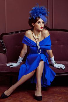 Old Fashioned retro great britain style photography. Beautiful young caucasian model in blue dress and fashion makeup and hat posing sitting on the artificial sofa and looking into the distance.