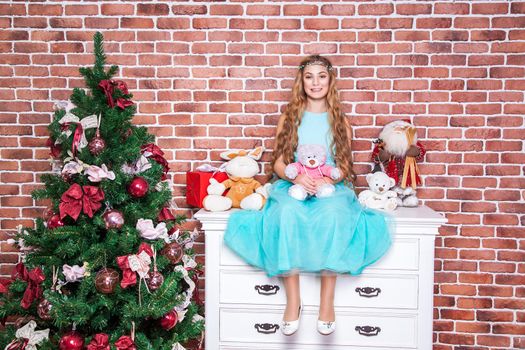 Cheerful teenage long haired blonde sit on a white nightstand near christmas tree, toothy smile and looking at camera. Studio shot