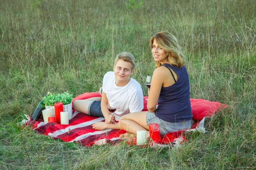 Beautiful young happy loving couple on picnic lying down on plaid in field on sunny summer day enjoying, holding and drinking wine and resting. looking at camera and smiling.