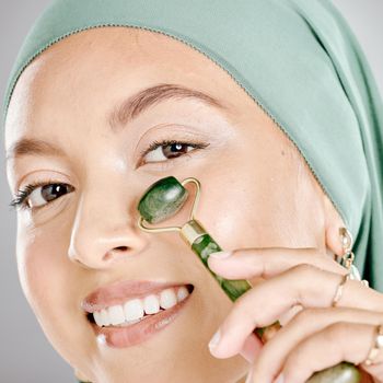 Muslim woman smiling and using a facial roller on her skin. Beauty model doing her morning skin care routine with a jade roller. Young female using a gua sha product for healthy smooth skin