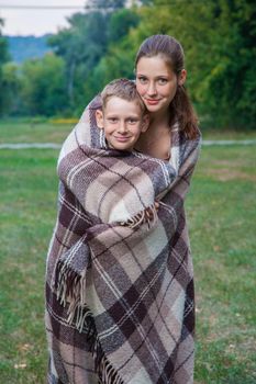 Young sister and brother with freckles on their faces stand covered with plaid in park, get warm, smiling and looking at camera.