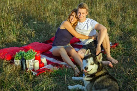 Beautiful young happy loving couple on picnic lying down on plaid with their dog in field on sunny summer day enjoying and resting. looking at camera and smiling.