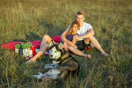 Beautiful young happy loving couple on picnic lying down on plaid with their dog in field on sunny summer day enjoying and resting. looking at camera and smiling.