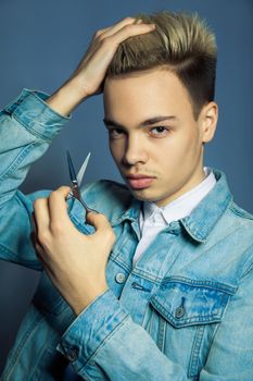 Portrait of fashion blonde Barber hairstylist with scissors in hand near eye on blue background.