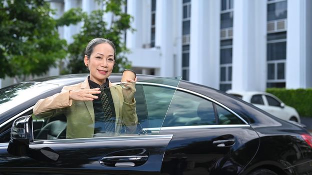 Confident mature business woman standing behind a car with opened door and smiling to camera.