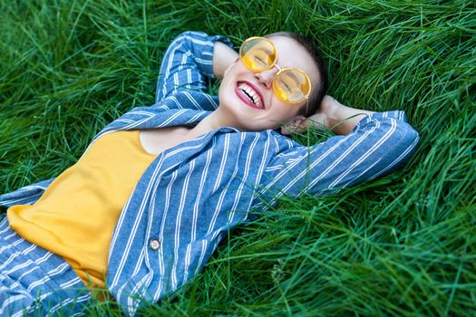 Portrait of Happy fancy young woman with short hair in casual blue striped suit, yellow shirt, glasses lying down on green grass, holding hands behind head, relaxing with toothy smile. summertime shot