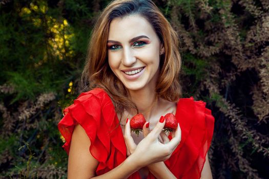 Young beautiful happy funny girl with red dress and makeup holding strawberry in summertime in the park. healthy lifestyle, diet beauty and happiness concept.