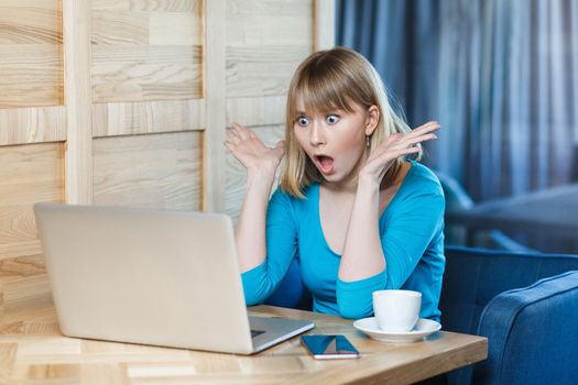 Side view portrait of emotional shocked young businesswoman in blue t-shirt are sitting in cafe, reading news and remotely working with suprised big eyes and raised arms looking to the laptop. Indoor