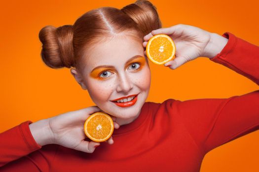 Young beautiful funny fashion model with orange slice on orange background. with orange makeup and hairstyle and freckles. studio shot, looking at camera with smile.