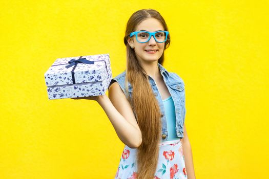 Portrait of a happy hipster smiling girl in dress and blue glasses, holding present box and toothy smile isolated over yellow background. Indoor studio shoot