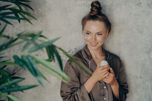 Image of caucasian cheerful woman in silk brown pajama smiling while applying face cream isolated over concrete wall background. Daily routine procedure for rejuvenation and care of skin