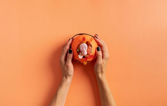 Happy Halloween concept. Scary woman hands with black nails holding pumpkin with sweets, flat lay on orange background
