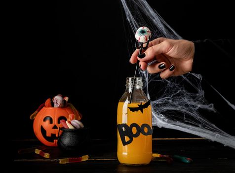 Happy Halloween concept. Woman hand with black nails holding scary colorful Halloween cocktail with party decorations on dark background
