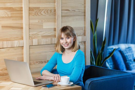 Portrait of beautiful satisfied young girl freelancer with blonde bob haircut hair in blue t-shirt are sitting in cafe and working on laptop with toothy smile and looking at camera. Indoor,