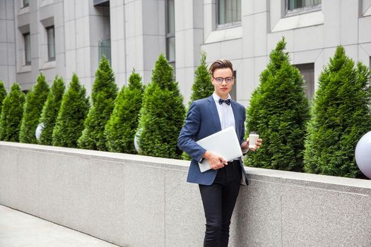 Attractive young businessman with laptop and coffee in hands on office building background. holding laptop and coffee and looking at camera.