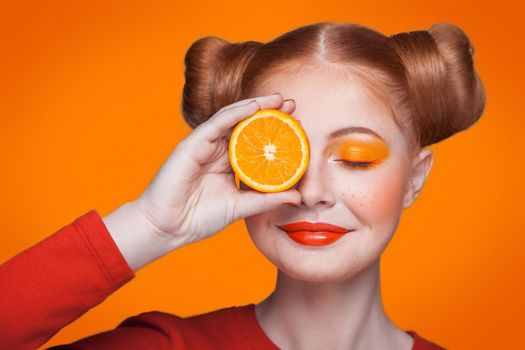Young beautiful funny fashion model with orange slice on orange background. with orange makeup and hairstyle and freckles. holding orange between eyes with nice smile.