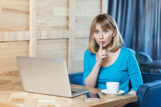 Be quiet please! Portrait of young girl freelancer with blonde hair in blue t-shirt are sitting in cafe and making video call on laptop, lean finger on mouth showing silence, looking at camera
